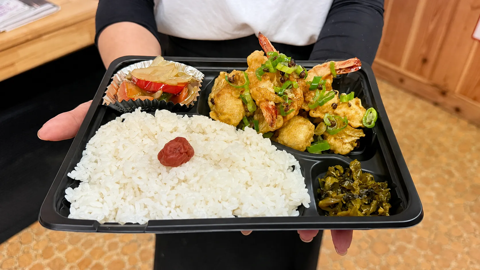 海老イカ天弁当を両手で持った様子。黒い容器に天ぷら、ご飯、野菜の餡かけ、高菜を盛り付け
